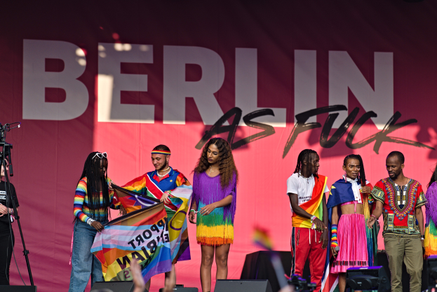Eine Gruppe von Menschen auf einer Bühne, mit einer Frau, die eine Fahne in der Mitte hält, einer Person mit einer Kamera links und einem Banner im Hintergrund mit der Aufschrift 'Berlin as Fuck 2019'.