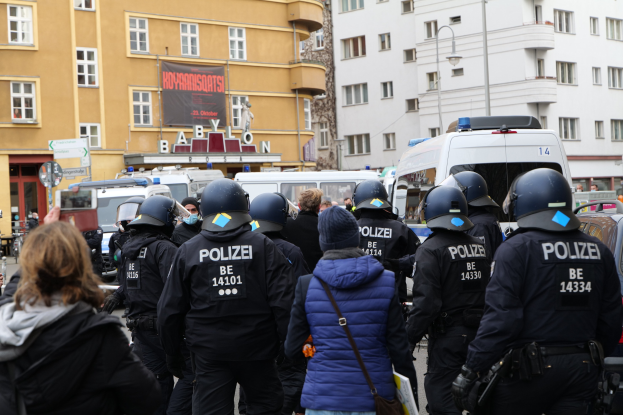 Polizeibeamte in Uniform vor einer Menge von Helmträgern während einer Demonstration in Berlin, mit Fahrzeugen, Gebäuden und einem Kameramann im Hintergrund.