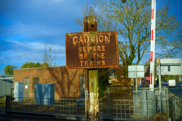 Vorsichtsschild an einem Bahnübergang mit Bäumen, Strommasten, einem Gebäude, Containern, Fahrzeugen und einem bewölkten Himmel.