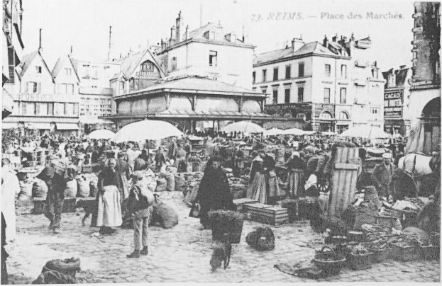Schwarz-weiß-Foto eines belebten Stadtmarkts mit Menschen, Schirmen, verstreuten Körben und Gebäuden im Hintergrund.
