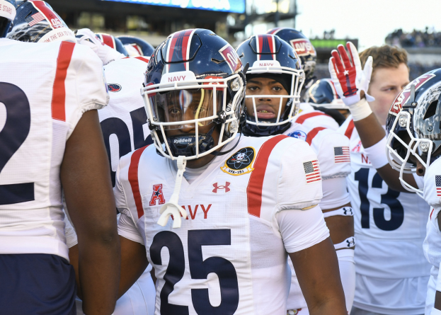 Gruppe von Männern in Football-Uniformen auf einem Feld mit einer unscharfen Menge, Anzeigetafel und Himmel im Hintergrund.