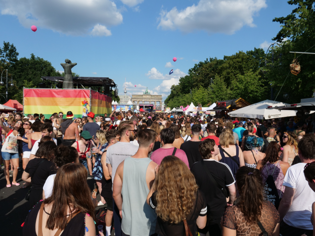 Eine große Menschenmenge, die eine Straße mit Zelten, Bäumen, Pfählen, Laternen und einer Statue gesäumt von Gebäuden im Hintergrund unter einem bewölkten Himmel mit Ballons während des Christopher Street Day in Berlin.