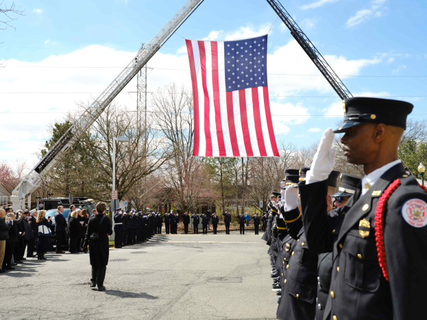 Gruppe uniformierter Personen, die vor einem Feuerwehrauto salutieren und eine amerikanische Flagge halten, mit Bäumen, Gebäuden und Laternen im Hintergrund.