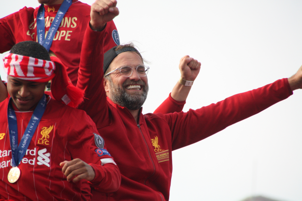 Liverpool-Trainer Jurgen Klopp feiert mit seinen Spielern den Sieg in der UEFA Champions League, umgeben von zwei medaillentragenden Personen und dem Himmel im Hintergrund.