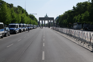 Eine Reihe von Polizeiwagen, die auf der Seite einer Straße vor dem Brandenburger Tor in Berlin, Deutschland geparkt sind, mit Menschen, die Fahrräder fahren und in der Nähe stehen, Barrieren, Bäumen und einem Bogen mit Statuen im Hintergrund.