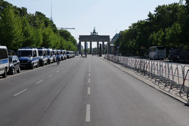Eine Reihe von Polizeiwagen, die auf der Seite einer Straße vor dem Brandenburger Tor in Berlin, Deutschland geparkt sind, mit Menschen, die Fahrräder fahren und in der Nähe stehen, Barrieren, Bäumen und einem Bogen mit Statuen im Hintergrund.