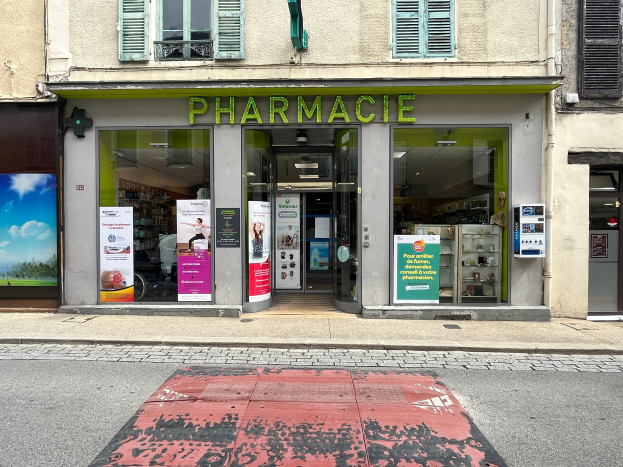 Eine Apotheke mit Glaswänden und -türen an einer Straßenecke, mit Postern an den Wänden und einer Straße im Vordergrund.