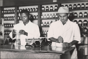 Zwei Männer in weißen Kitteln und Hüten stehen an einem Tresen in einer Apotheke mit einer Waage und einer Schüssel, mit Regalen voller Flaschen im Hintergrund, dargestellt in Schwarz-Weiß.