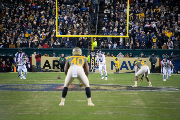 Ein Fußballspiel wird in einem Stadion mit Spielern in Sportkleidung und Helmen im Vordergrund und einer Zuschauermenge im Hintergrund sowie sichtbaren Pfählen und Tafeln mit Text gespielt.