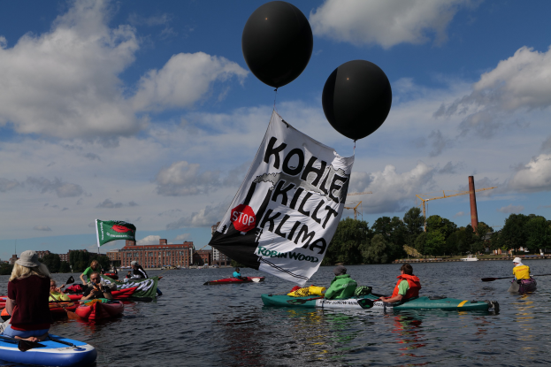 Gruppe von Menschen in Kajaks, die auf dem Wasser paddeln, mit einem Banner, auf dem "Kohle Kill Klima" steht, und Bäumen, Gebäuden und Kränen im Hintergrund unter einem klaren blauen Himmel.