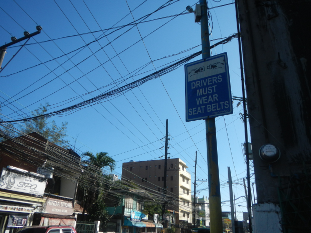 Stadtstraße mit fahrenden Autos, Strommäste mit Drähten, Gebäude, Bäume und Schildern, mit einem "Fahrer müssen den Gurt anlegen"-Schild an einem Strommäst.