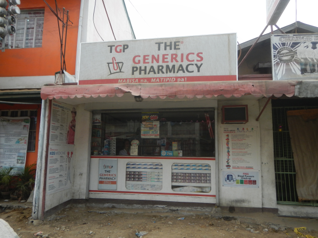 Eine Apotheke namens "Die Generika Apotheke" ist von Gebäuden, Strommasten, Kabeln, Werbetafeln, Topfpflanzen und Kraftfahrzeugen auf der Straße unter einem klaren blauen Himmel umgeben.
