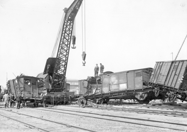 Schwarß-weißes Bild eines Zugunglücks mit einem Kran, der ein Zugwaggon auf die Schienen hievt, mehrere Personen in der Nähe, Gebäude und einen klaren Himmel im Hintergrund.
