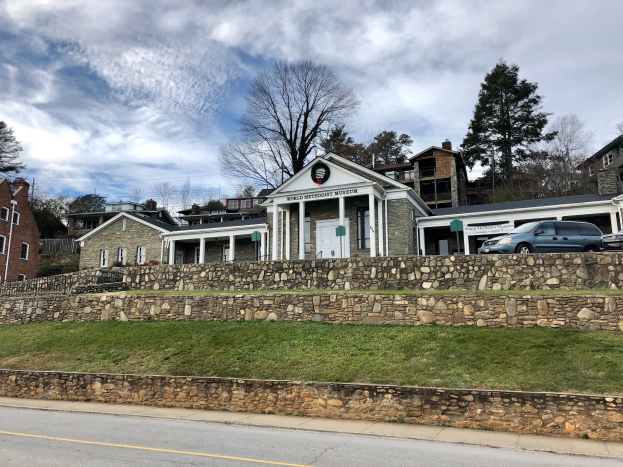 Steinwand mit geparkten Autos, benachbarten Gebäuden mit Fenstern und Türen, Bäumen und einem bewölkten Himmel im Hintergrund; das Weltkulturerbe-Museum, kürzlich wiedereröffnet.