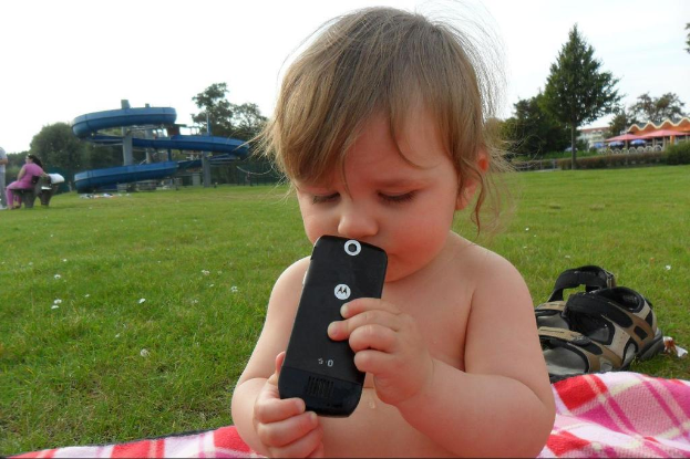 Ein kleines Baby hält ein Mobile, während es auf einem karierten Handtuch auf einer grünen Wiese sitzt, mit Bäumen, einer Bank, einer Dame auf der Bank und einem Reiter im Hintergrund.