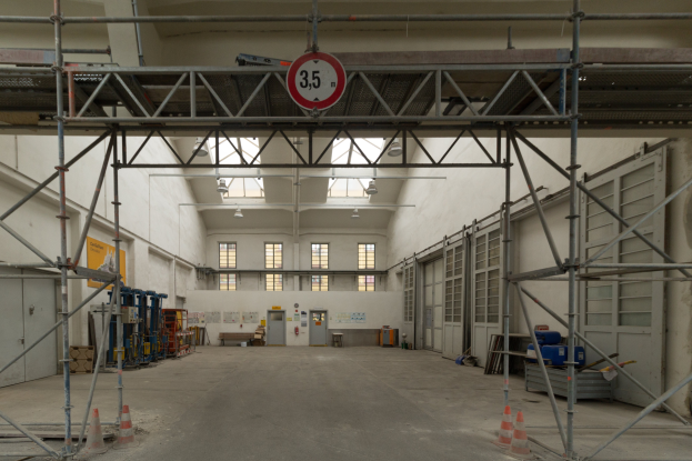 Ein großes Lager mit umfangreichen Gerüsten, Verkehrskegeln auf dem Boden, Metallstangen, einem Schild, verschiedenen Maschinen, einer Tür, Fenstern, einer Wand und einem Dach mit Deckenleuchten.