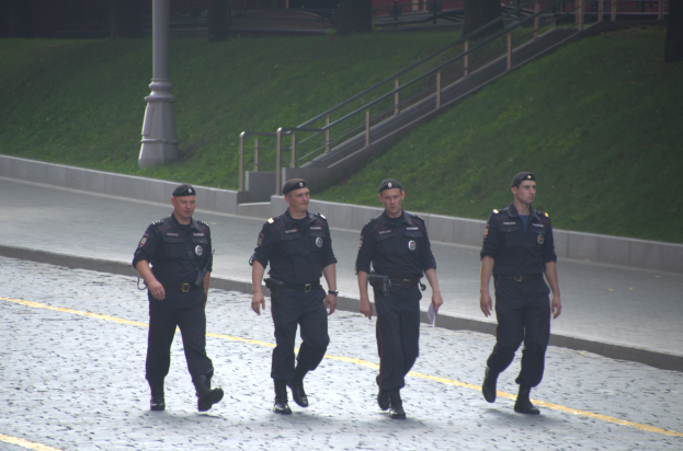 Vier Polizisten in Uniform gehen eine Straße entlang und halten Papiere in den Händen, mit einem Gebäude, Bäumen und einem Pfahl im Hintergrund.
