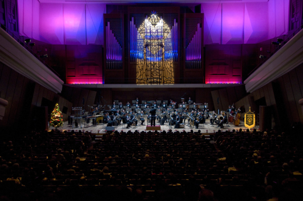 Großer Saal mit Publikum bei einem Konzert; Musiker auf der Bühne, einige sitzend, mit einem Weihnachtsbaum und geschmückten Wänden im Hintergrund.