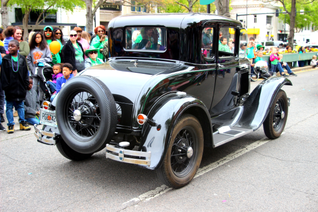 Ein Oldtimer fährt an einer Menge von Menschen vorbei, die Hüte und Schutzbrillen tragen, mit Bäumen, Gebäuden, Laternen und Texttafeln im Hintergrund.