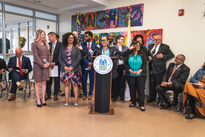 Eine Gruppe von Menschen, die vor einem Podium mit Mikrofon und einer Tafel mit der Aufschrift "Sancho City Councilman Joins City Council" stehen und sitzen, mit Flaggen, Feuerlöscher und Deckenlampen an der Rückwand.