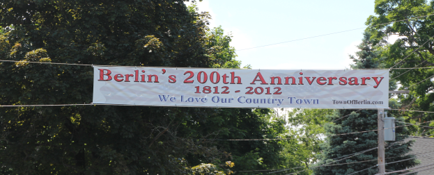 Ein Banner mit der Aufschrift 'Berliner Jubiläum 200' hängt an einem Draht in der Vordergrund, mit Bäumen im Hintergrund und einem Haus auf der rechten Seite, unter einem sichtbaren Himmel.