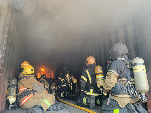 Feuerwehrleute in Helmen und Sauerstoffflaschen sitzen im Heck eines raucherfüllten Trucks, während im Hintergrund ein großes Feuer brennt.