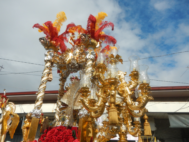 Ein großes goldenes und rotes schwimmendes Gebilde, geschmückt mit Blumen und anderen Dekorationsgegenständen bei einem Karnevalsumzug, mit einem Gebäude, Strommasten mit Drähten und einem bewölkten Himmel im Hintergrund.