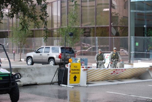 Eine Gruppe von Polizeibeamten in Uniform steht vor einem Gebäude mit Glaswänden, mit Fahrzeugen im Vordergrund, Bäumen und einem Zaun im Hintergrund und einem Wasserzeichen im rechten unteren Eck.