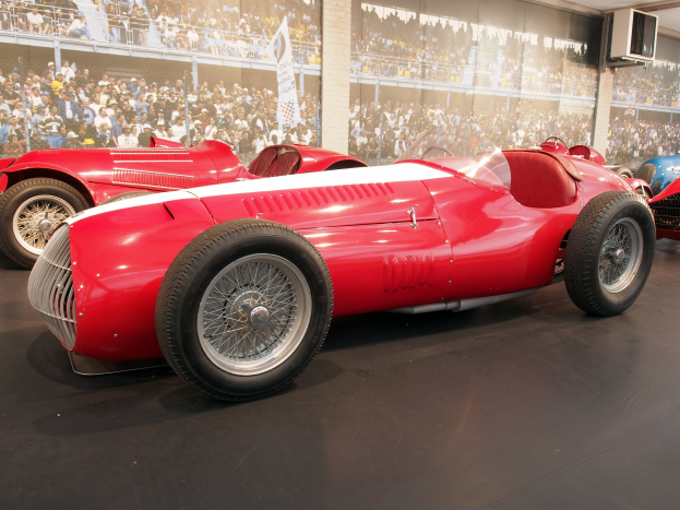 Ein roter Rennwagen in einem Museum ausgestellt, umgeben von anderen Autos, mit einer großen Menge, Bannern und Deckenleuchten im Hintergrund.