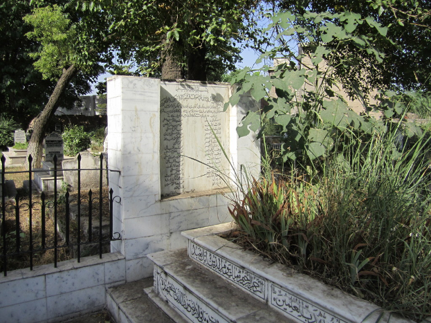 Ein Gedenkstein neben einem Geländer in einem Friedhof, mit Treppen unten, Gras und Bäumen rechts, Gräbern dahinter und Gebäuden und Bäumen im Hintergrund.