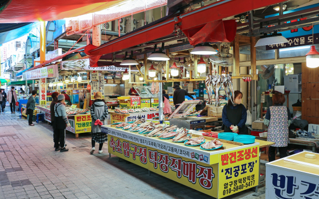Ein belebter Straßenmarkt in Seoul, Südkorea, mit Menschen, Tischen mit verschiedenen Gegenständen und Gebäuden im Hintergrund unter einem klaren blauen Himmel.