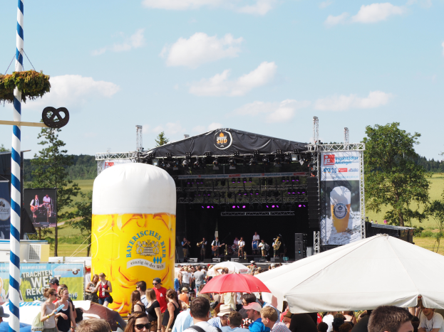 Eine große Menschenmenge beim Oktoberfest in München, Deutschland, viele tragen Kappen und Goggles, einige halten Schirme, ein Zelt im Vordergrund, ein dekorierter Pfahl links, Bäume und ein bewölkter Himmel im Hintergrund und eine Bühne mit einem Banner und Musikern.