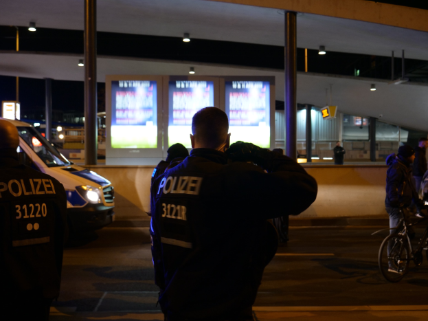 Gruppe von Polizeibeamten vor einem Gebäude bei Nacht mit einem geparkten Auto links und einem Radfahrer rechts, beleuchtet von Bildschirmen und Lichtern im Hintergrund.