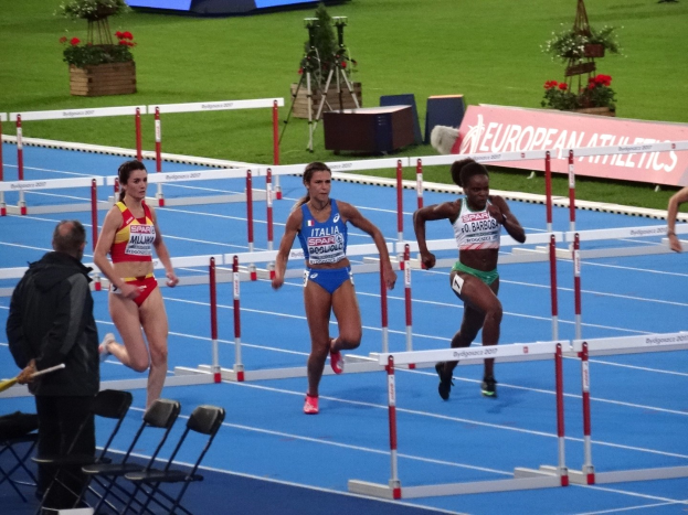 Frauenstaffel über Hürden auf einem Rasenplatz mit einer Stabhalterin links im Bild, Stühle und ein Schreibtisch im Vordergrund, Blumentöpfe und Texttafeln im Hintergrund.