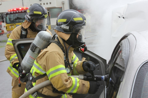 Feuerwehrleute in Schutzausrüstung löschen ein Autofeuer mit einem Schlauch, wobei Rauch aus dem Fahrzeug und anderen Fahrzeugen und einem Gebäude im Hintergrund sichtbar ist.