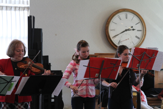 Drei Frauen auf der Bühne mit Violinen spielend, mit Notenpulten und Notenblättern davor, während ein Zuschauer im Vordergrund und eine Uhr an der weißen Wand im Hintergrund sowie ein weißer Vorhang in der linken Ecke zu sehen sind.