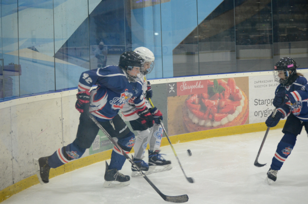Gruppe junger Menschen, die Eis Hockey auf einer Indoor-Eisfläche spielen, mit Helmen, Sportbekleidung und Hockey-Schlägern, sowie einem Plakat im Hintergrund auf einer Glaswand.