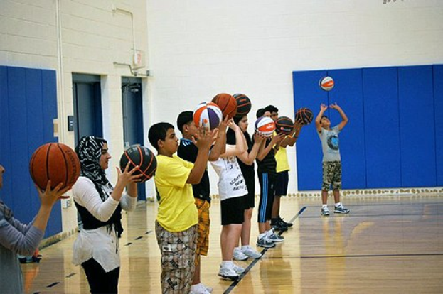 Junge Menschen mit Basketballs auf einem Basketballfeld während eines Camps, mit Türen und einer Wand im Hintergrund.