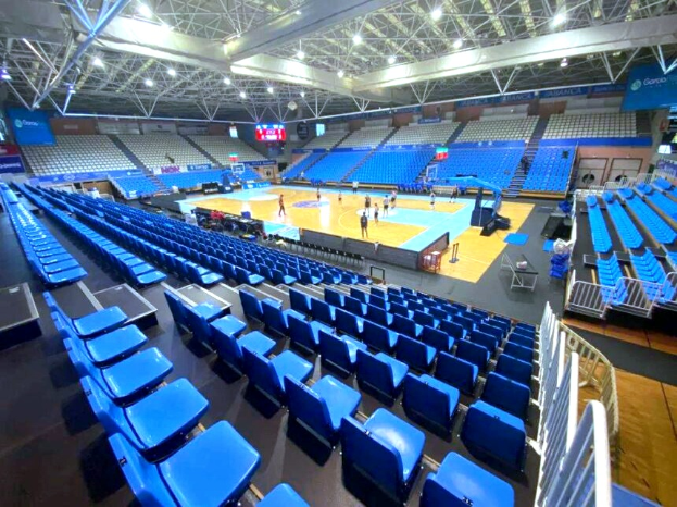 Ein großes Hallenstadion in Sarajevo, Bosnien und Herzegowina, mit blauen Stühlen, einem Basketballfeld und ein paar Menschen auf dem Feld, unter einer Decke mit Lampen und Trägern.