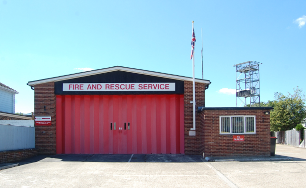 Feuerwehr- und Rettungsdienstgebäude mit roter Tür, Fenstern, einem Namensschild, einer Fahnenstange mit einer Fahne, einem Metallturm, einem Zaun, einer Gruppe von Bäumen und einem bewölkten Himmel.