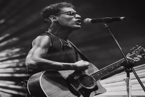 Ein Mann mit Brille spielt eine Gitarre vor einem Mikrofon, performt in der O2 Academy Brixton, dargestellt in Schwarz-Weiß.