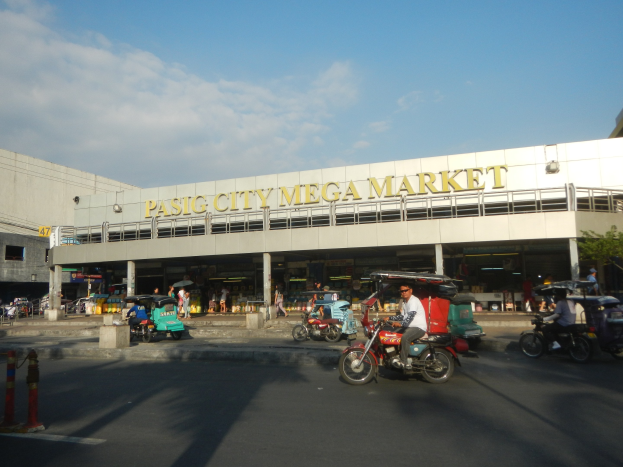 Eine belebte Straßenszene am Pasig City Mega Market mit Fahrzeugen und Fußgängern, ein Gebäude mit Text im Hintergrund und eine bewölkte Himmelslandschaft.