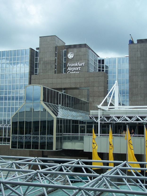Ein großes, modernes Flughafengebäude mit Glaswänden und Text darauf, umgeben von gelben Flaggen und Eisenstangen, unter einem Himmel voller weißer, flauschiger Wolken.