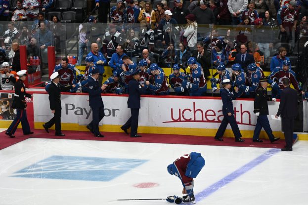 Ein Hockey-Spieler auf dem Eis mit einem Stock in der Hand, umgeben von einer Gruppe von Menschen mit Mützen und Hockey-Stöcken, mit Zuschauern und Brettern, auf denen "Colorado Avalanche vs San Jose Sharks - 3/24/15 NHL Pick, Odds, and Prediction" steht.
