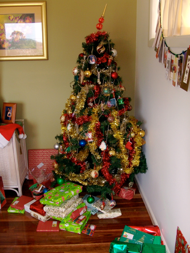 Ein geschmückter Weihnachtsbaum auf der rechten Seite mit Geschenken am Boden, ein Photo Rahmen an der linken Wand und ein Vorhang am rechten Fenster.