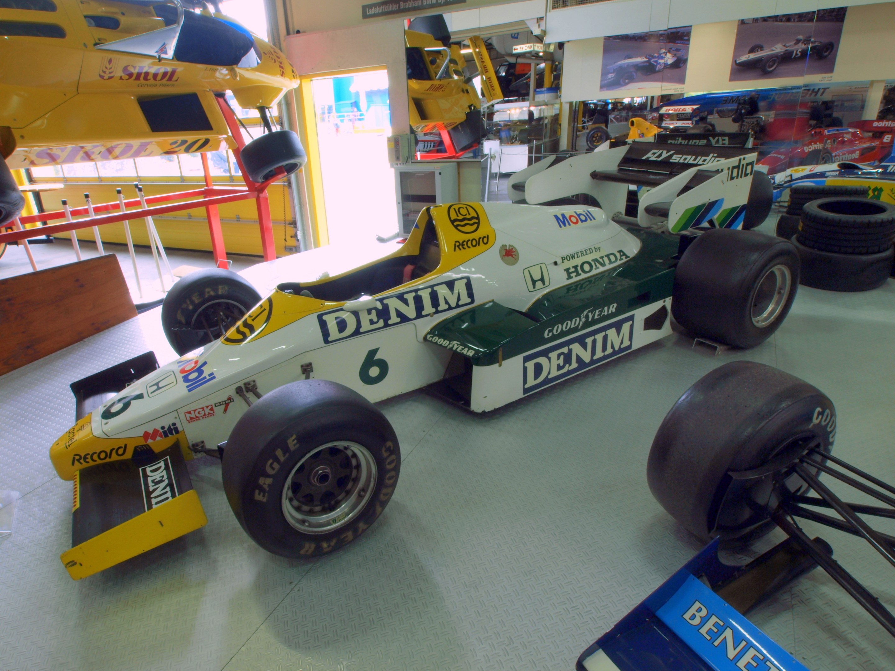 Eine unordentliche Garage mit verschiedenen Rennwagen, Reifen, einem hölzernen Objekt, einem Geländer, einer Glastür, gerahmten Fotos an der Wand und Deckenbeleuchtung.