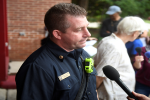 Ein Polizeibeamter in Uniform spricht mit Reportern, die ein Mikrofon halten, während andere in der Nähe stehen, vor einer Backsteinwand und Bäumen.