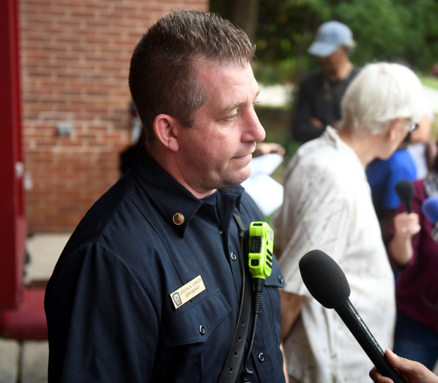 Ein Polizeibeamter in Uniform spricht mit Reportern, die ein Mikrofon halten, während andere in der Nähe stehen, vor einer Backsteinwand und Bäumen.