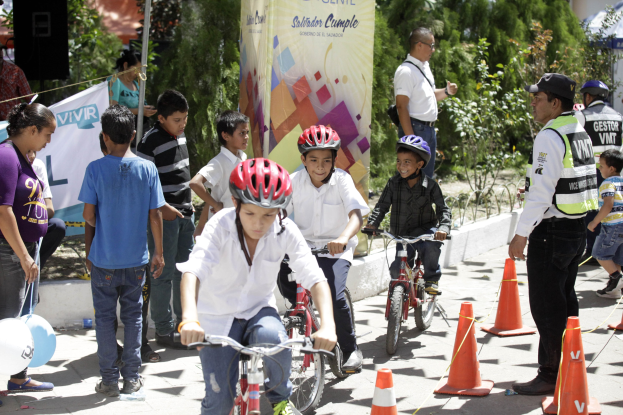 Eine Gruppe von Kindern, die auf Fahrrädern eine Straße mit Verkehrskegeln hinunterfahren, wobei einige Helme tragen und andere in der Nähe stehen, mit einem Banner, Bäumen und Gebäuden im Hintergrund.