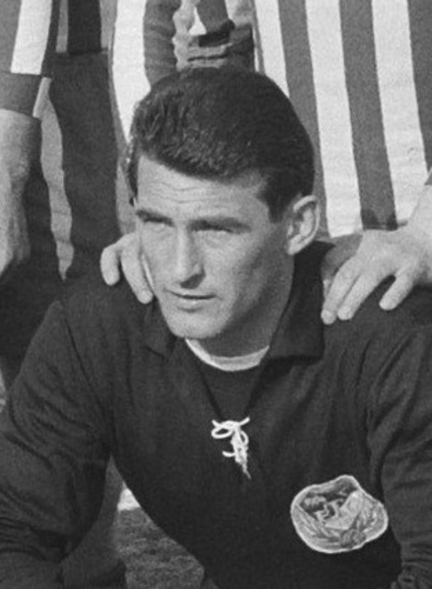 Ein Schwarz-Weiß-Foto eines Mannes, der auf einem grünen Fussballfeld sitzt und ein T-Shirt sowie eine Kette trägt, mit zwei Personen hinter ihm.